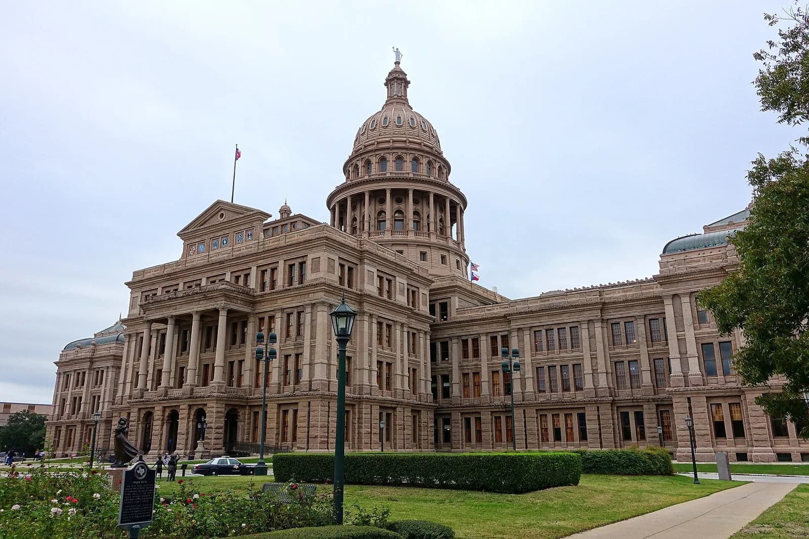 A large, ornate stone building with a prominent dome and columns stands under an overcast sky. Green lawns, rose bushes, and a statue are visible in the foreground.