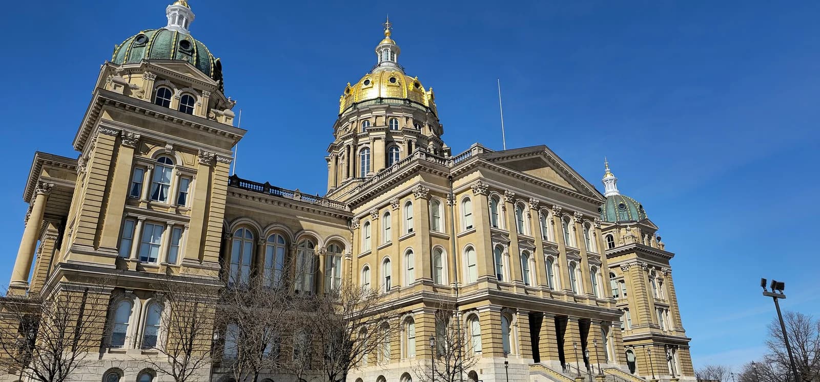 A large, ornate building with a central golden dome and two smaller green domes stands against a clear blue sky. The building features classical architectural elements, including columns, arched windows, and decorative cornices.