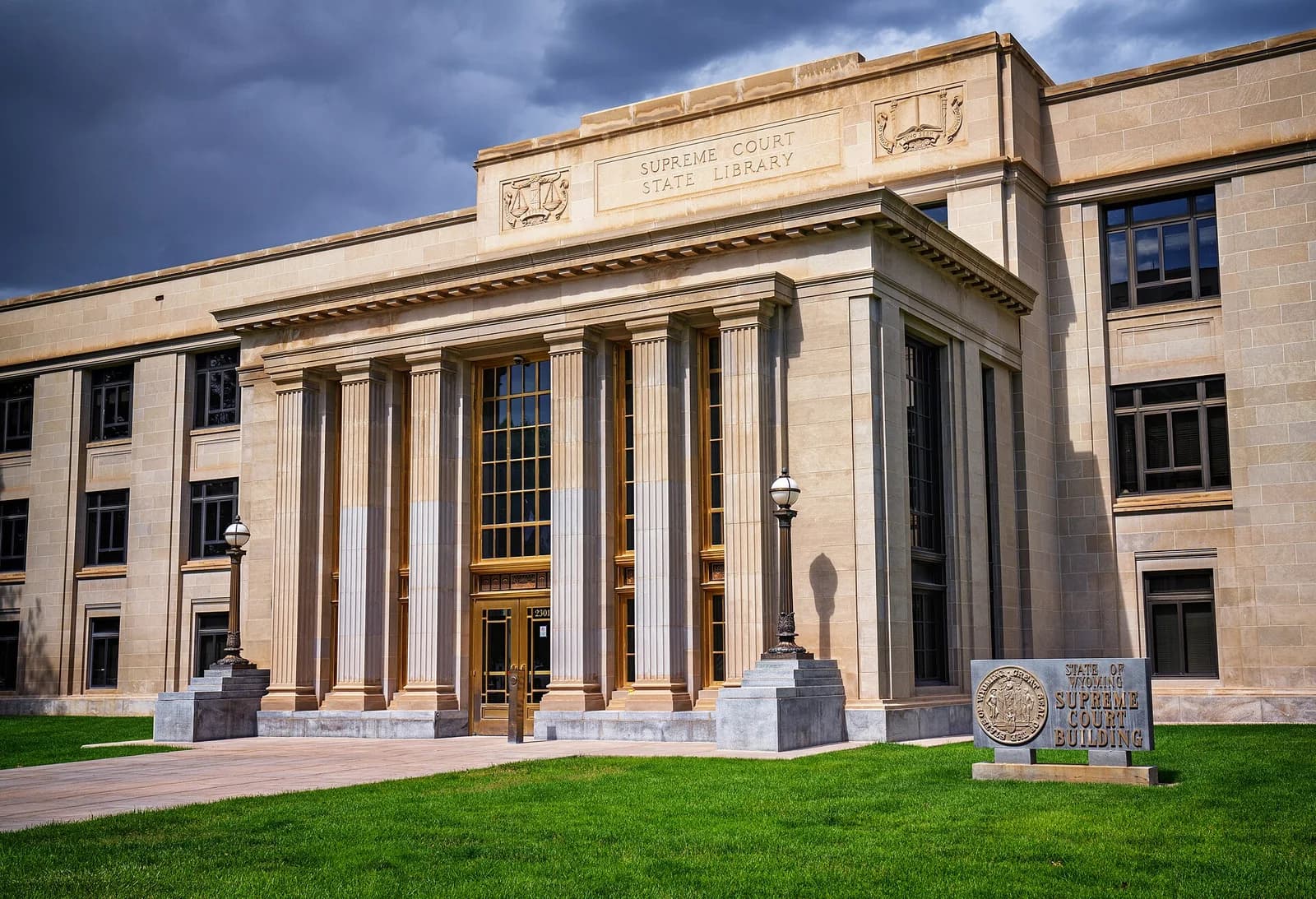 The Wyoming Supreme Court building, a large stone structure with columns and tall windows, stands under a dramatic cloudy sky. A sign for the "State of Wyoming Supreme Court Building" is visible in the foreground on a patch of green grass.