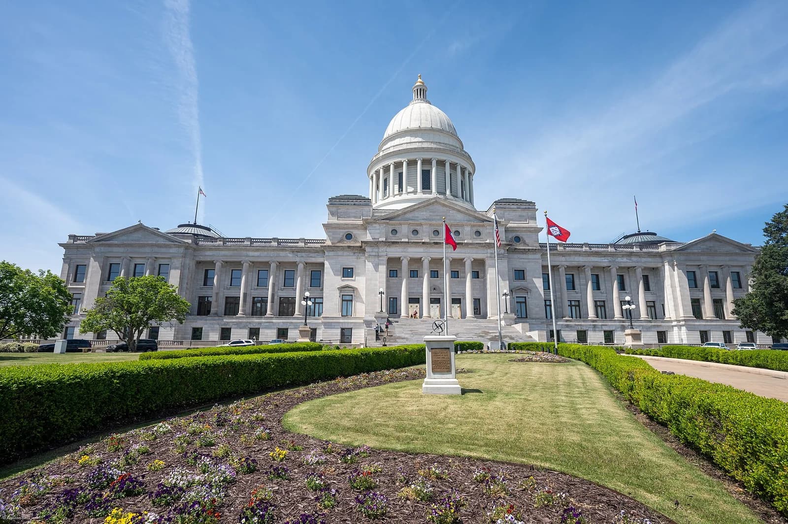 A grand, white stone building with a large central dome and columns stands under a clear blue sky. In the foreground, a manicured lawn with flower beds and hedges leads up to the building, with flags flying near its entrance.