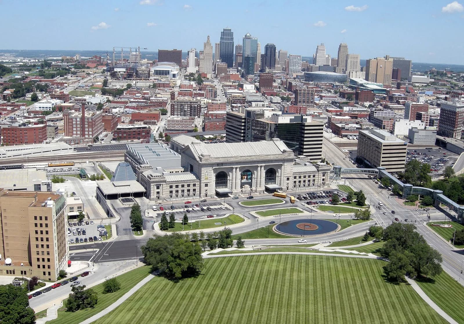 An aerial view shows a large, ornate stone building with a grand entrance and columns, surrounded by roads, parking lots, and a manicured green lawn. In the background, a dense cityscape with numerous high-rise buildings extends under a clear blue sky.