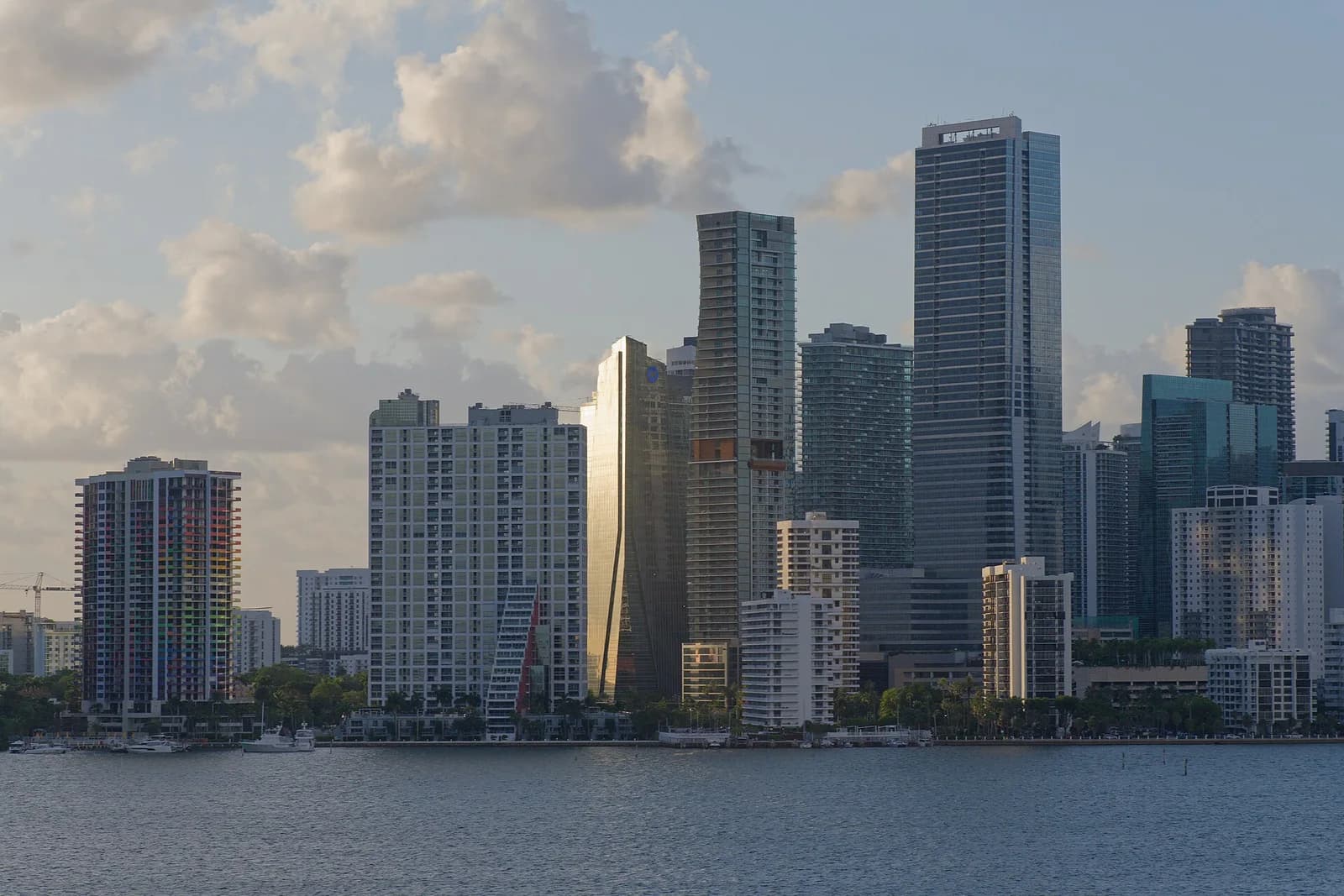 A wide shot of the Miami, Florida skyline from across the water, showing numerous high-rise buildings under a partly cloudy sky. The water in the foreground reflects the light from the sky.