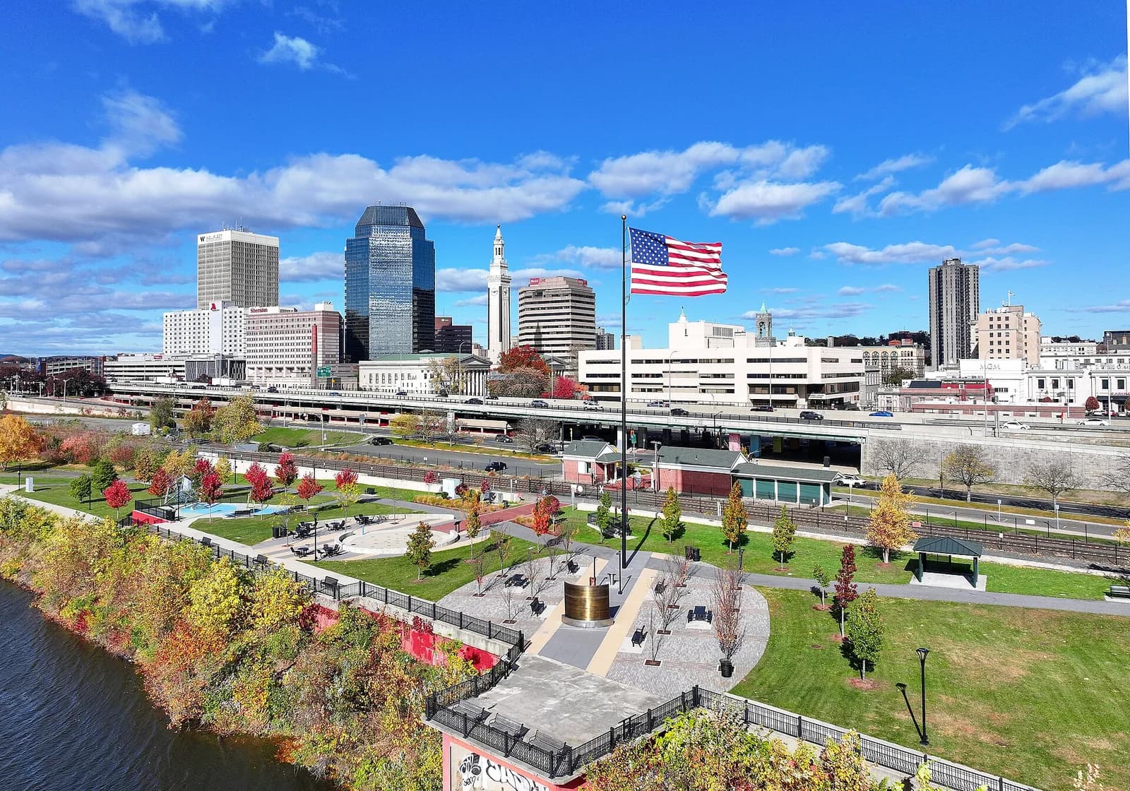 An aerial view of a city skyline with various buildings, including skyscrapers, under a blue sky with white clouds. In the foreground, a park with autumn trees and a large American flag on a flagpole overlooks a river and a highway.