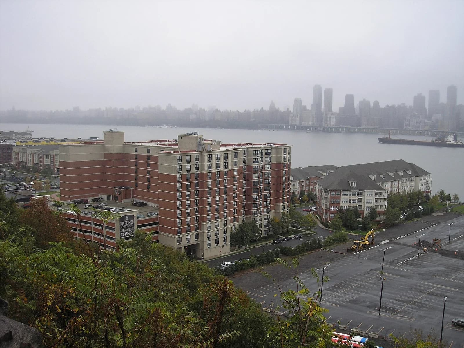 A cityscape view on a foggy day shows a river with a distant skyline and bridge, with several residential buildings and a large empty parking lot in the foreground. A promotional banner is visible on a parking garage.