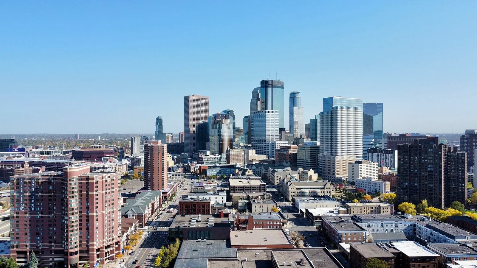 An aerial view of a city skyline under a clear blue sky, featuring a mix of modern skyscrapers and older, lower-rise buildings. A wide street with cars and trees runs through the foreground, leading towards the dense urban core.