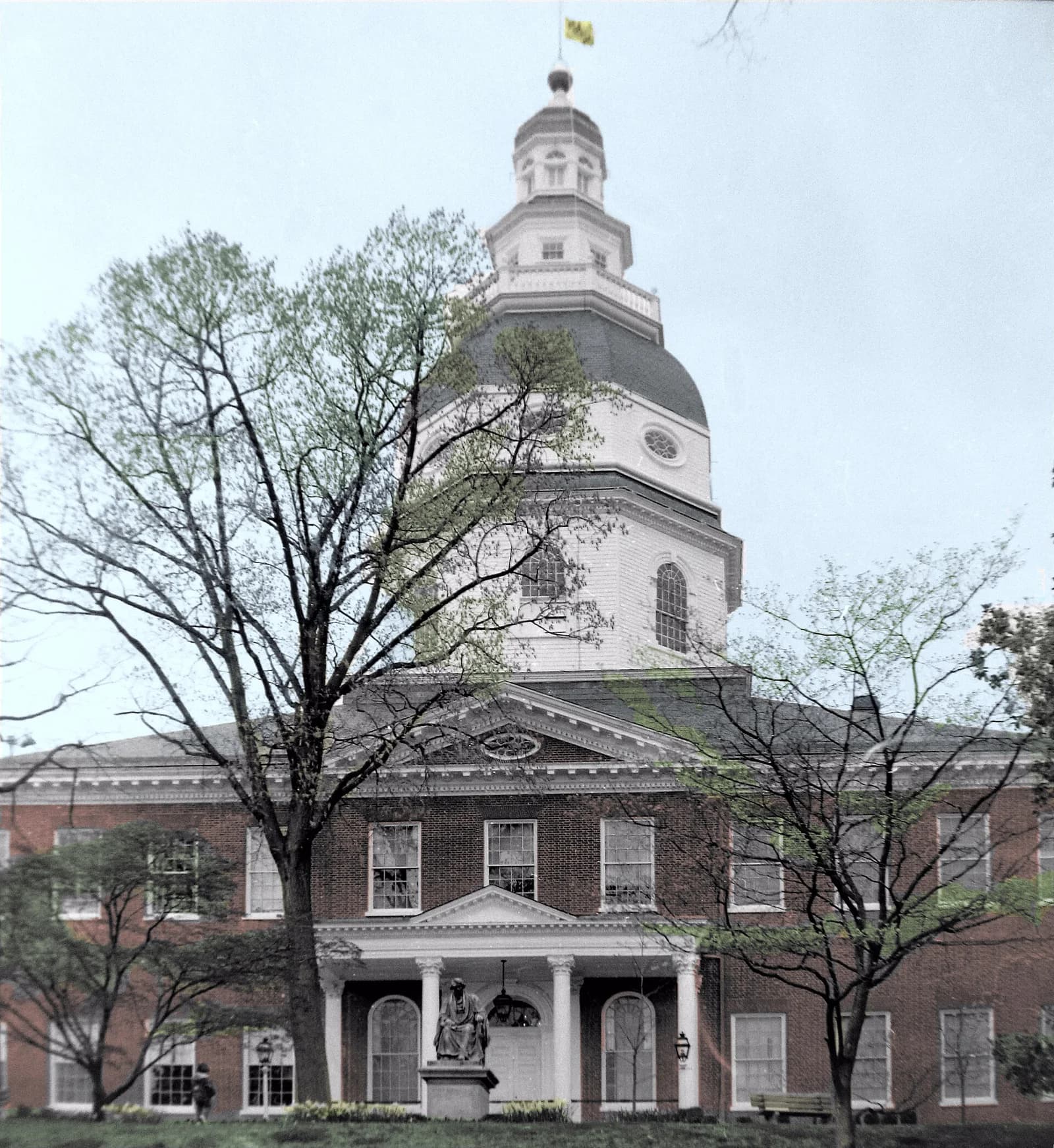 A brick building with a white dome and cupola is partially obscured by trees with new spring leaves. A statue of a seated figure is visible in front of the building's entrance, which features white columns.