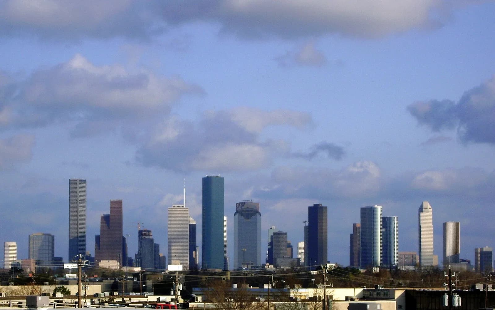 A wide shot of a city skyline shows numerous tall buildings under a partly cloudy blue sky. The foreground features rooftops and utility poles, with the cityscape rising behind them.