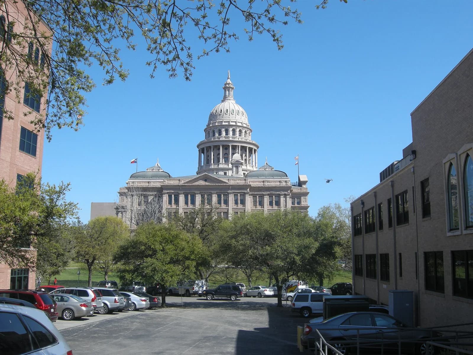 A large, domed institutional building with a flag flying on its roof stands prominently in the background, framed by trees and other buildings. In the foreground, a parking lot with several cars is visible, with a brick building on the right and a taller building on the left.