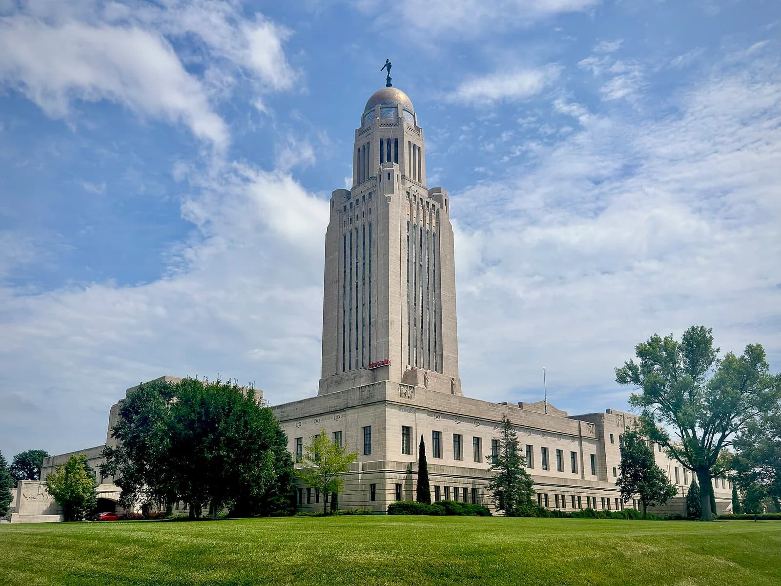 A tall, light-colored stone building with a prominent central tower topped by a golden dome and a statue stands against a partly cloudy blue sky. The building is surrounded by green lawns and several trees.