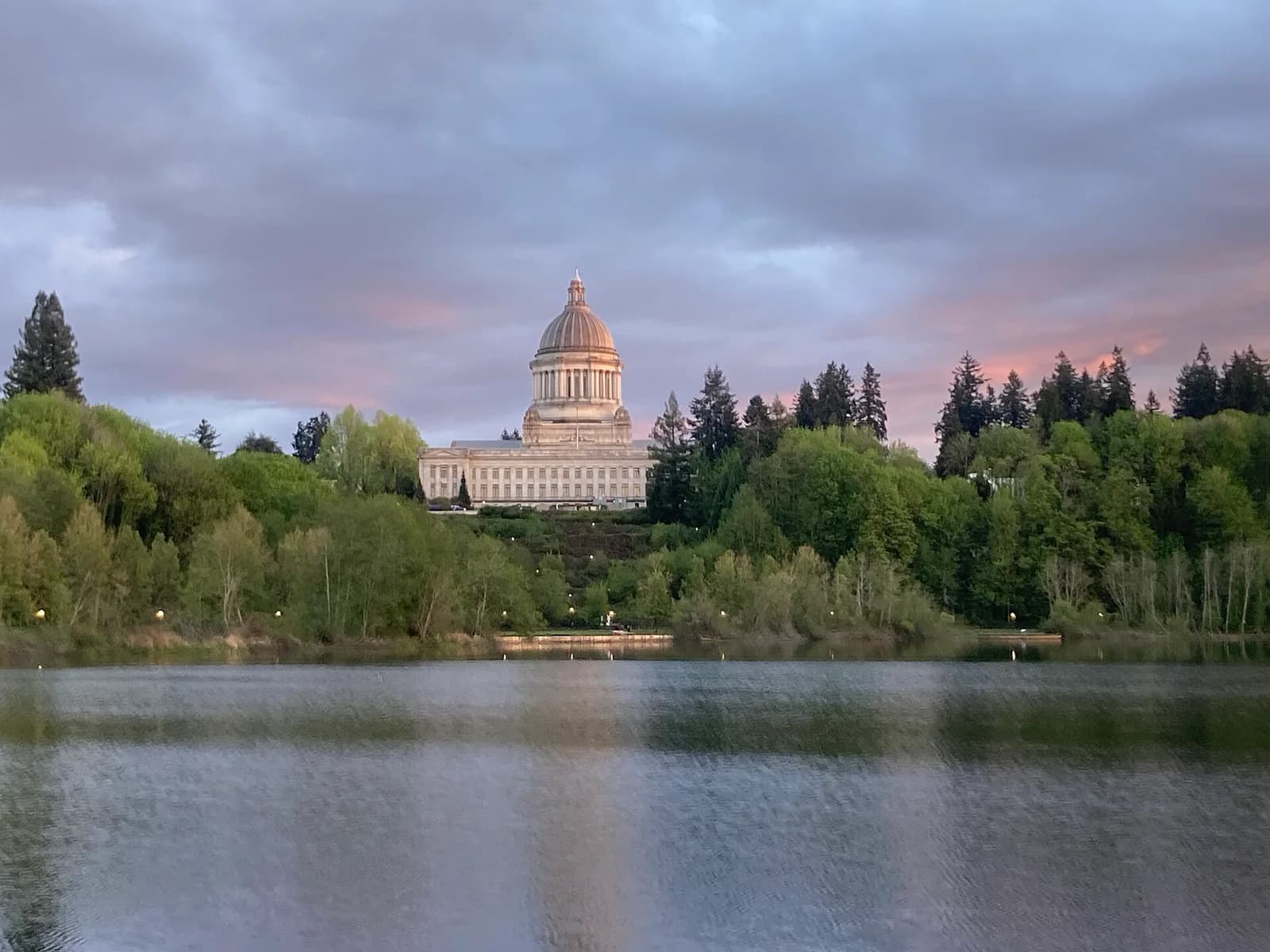 A large domed building with columns stands on a hill surrounded by green trees, overlooking a body of water. The sky above is cloudy with hints of pink and orange from a sunset.