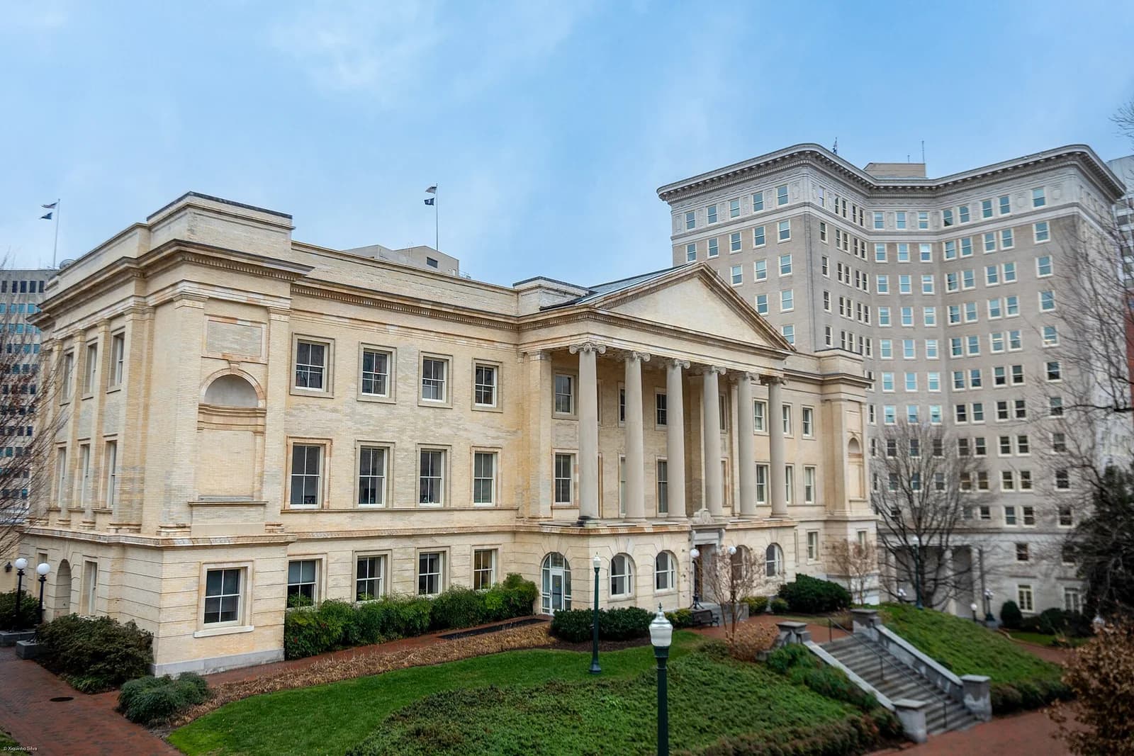 A large, light-colored neoclassical building with a prominent portico and columns stands on a grassy hill, with a taller, curved building in the background. Steps lead up the hill to the building, and bare trees are visible on the right.