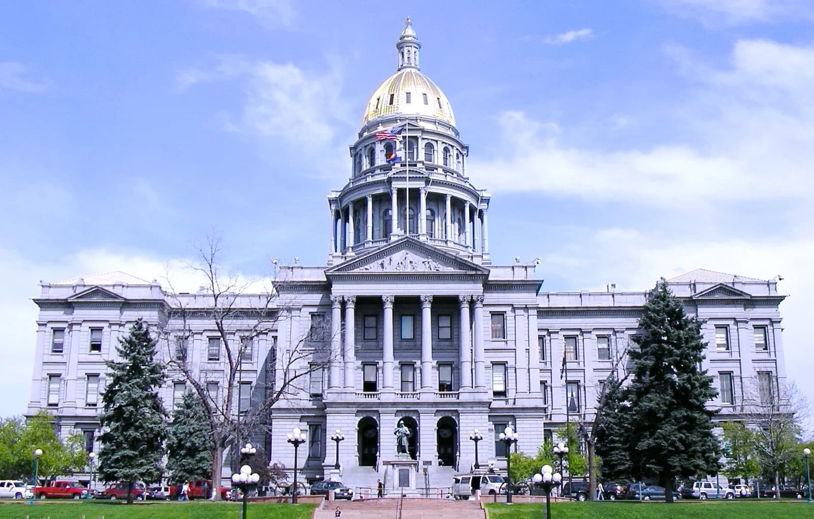 A large, ornate building with a golden dome and columns stands under a blue sky with scattered clouds. Green lawns and trees are in the foreground, with a few cars and distant people visible.