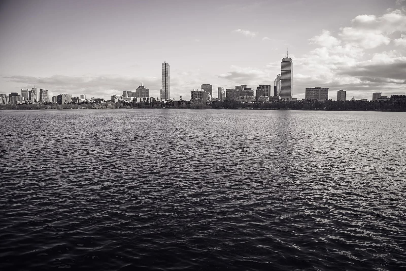 A black and white cityscape shows a skyline of buildings across a body of water, with a cloudy sky above. The water in the foreground is rippled, reflecting some light.