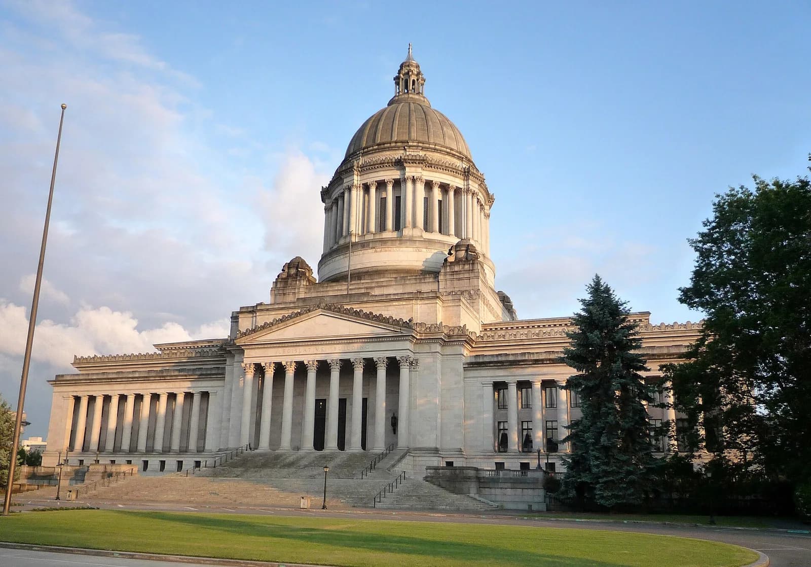 A large, neoclassical building with a prominent dome and columns stands under a partly cloudy sky. A wide lawn and trees are visible in the foreground and to the right of the building.