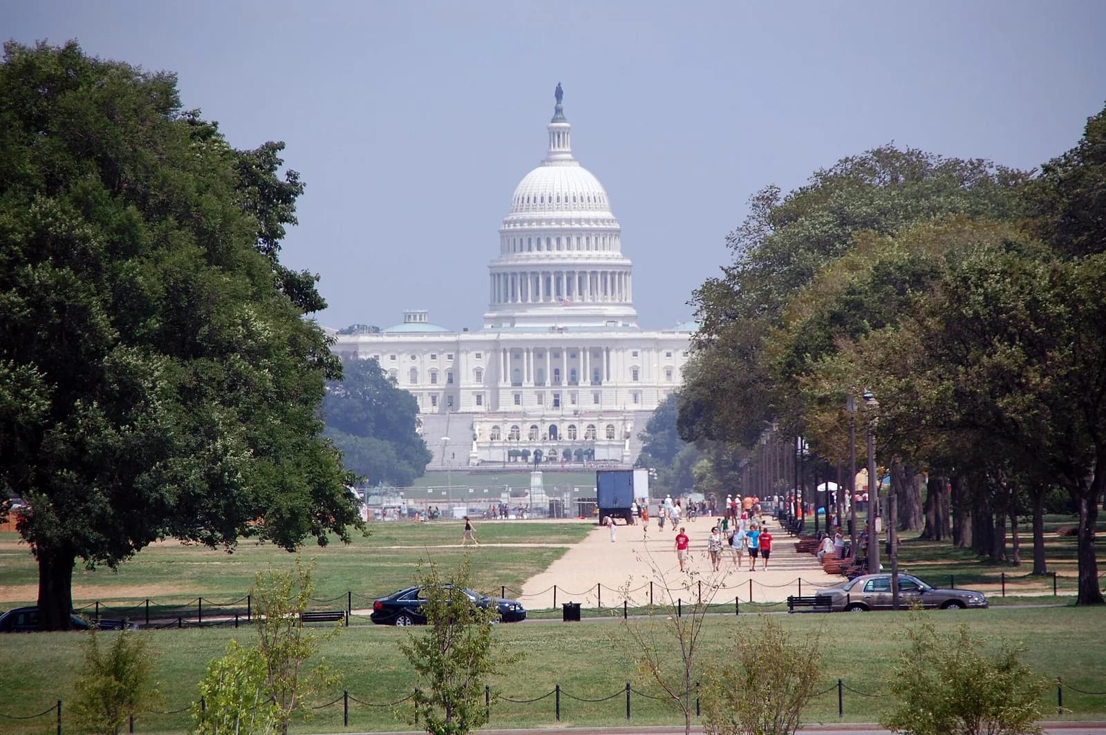 A large white domed building with columns is visible in the distance, framed by green trees on either side. A wide dirt path with people walking on it leads towards the building, with grassy areas and more trees in the foreground.