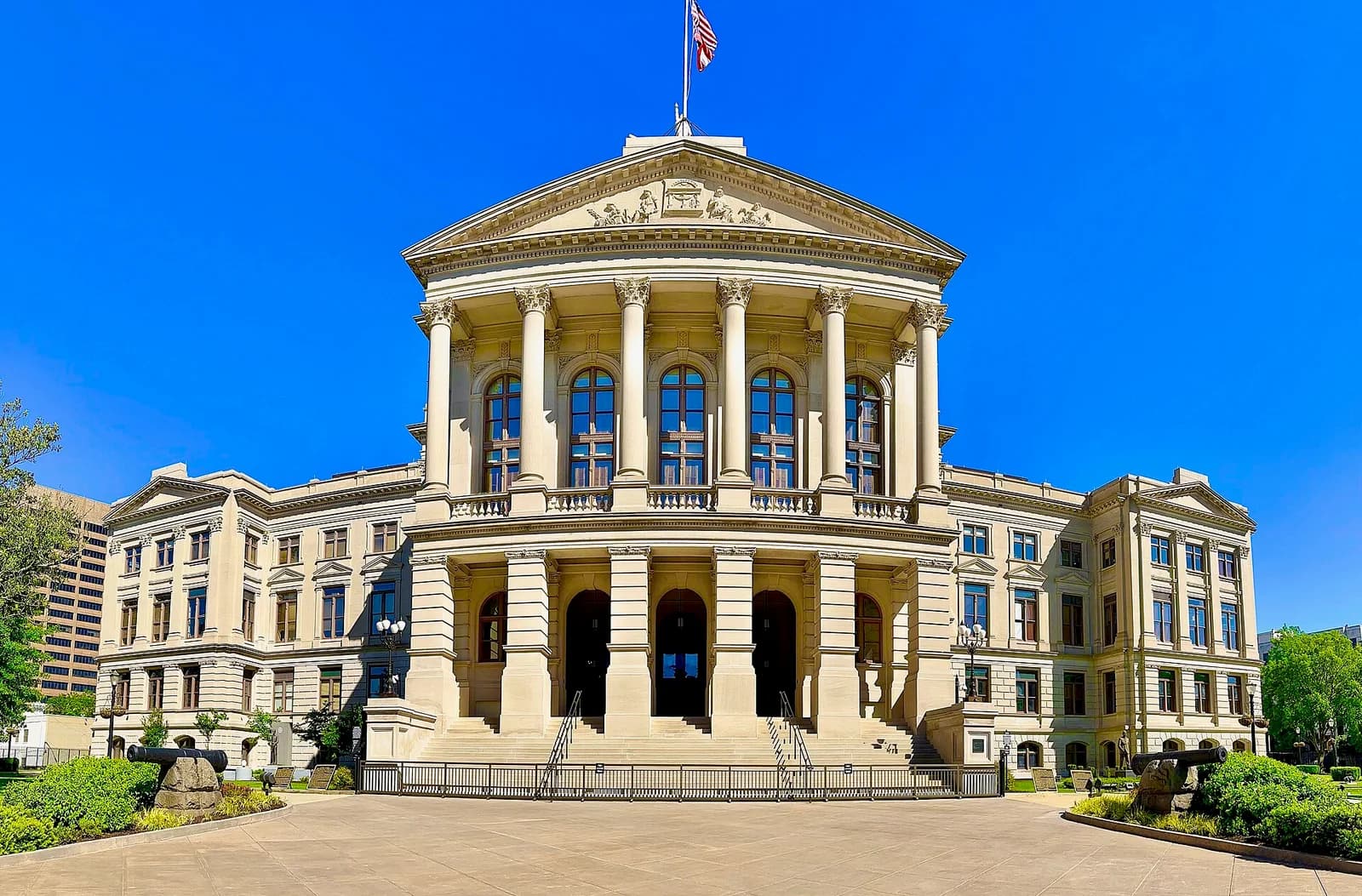 A grand, light-colored neoclassical building with a large portico and dome is visible under a clear blue sky. Cannons are visible on the lawn in front of the building.