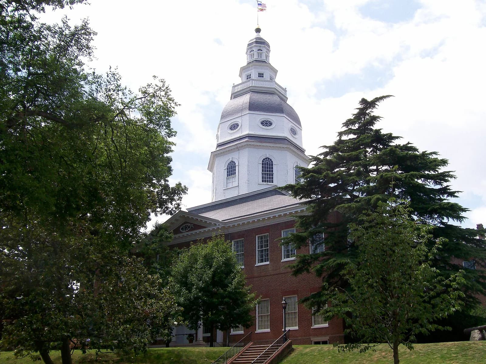 A red brick building with a prominent white dome and cupola is partially obscured by green trees. A set of stairs leads up to the building's entrance.