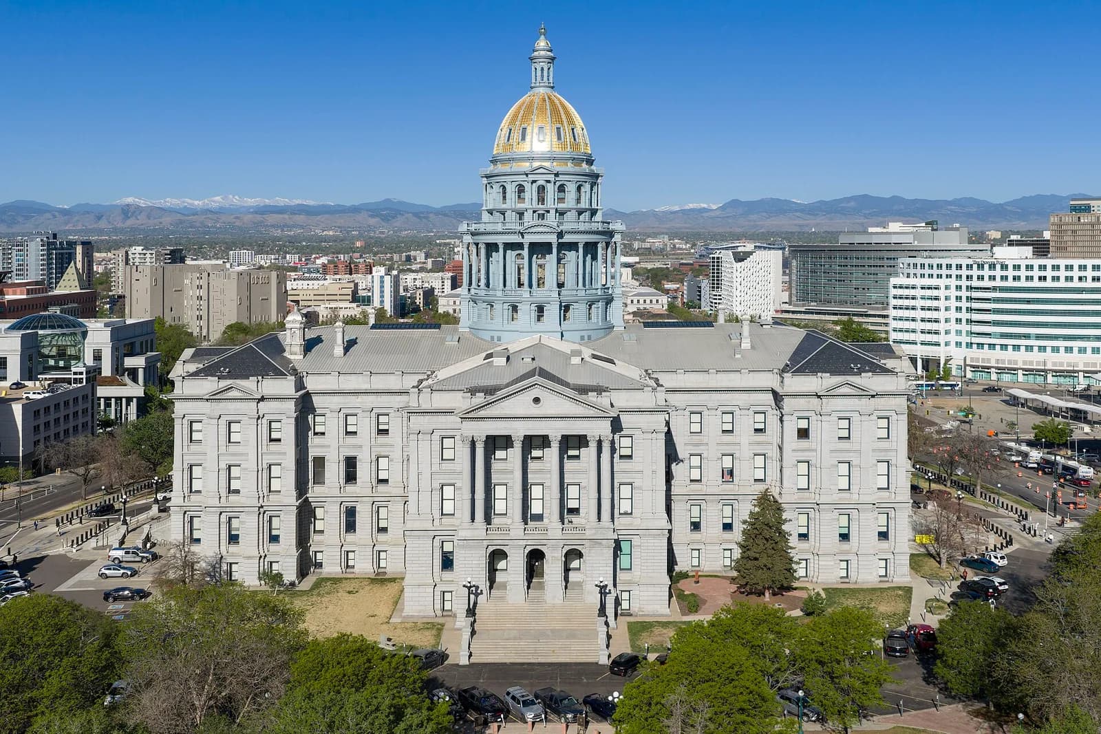 An aerial view shows a large, light-colored stone building with a prominent gold dome and a blue-gray cupola, surrounded by green trees and other city buildings. Snow-capped mountains are visible in the distant background under a clear blue sky.