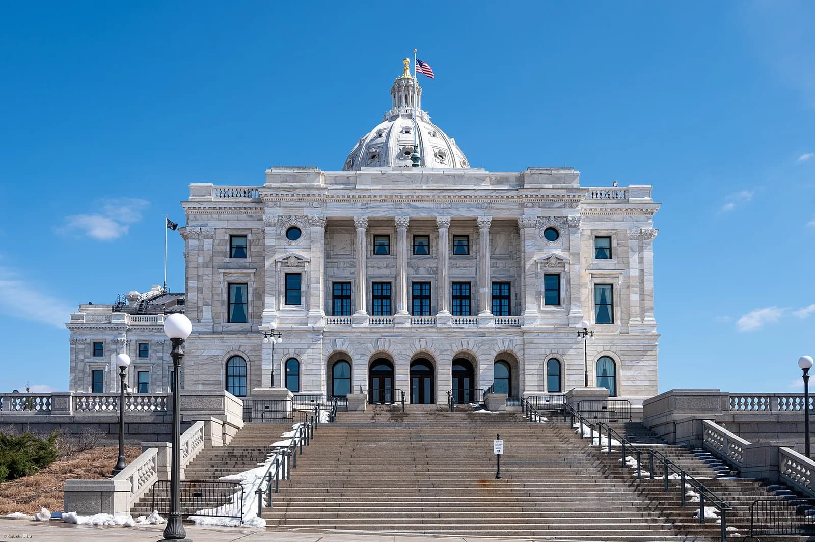 A grand, light-colored stone building with a large dome and an American flag on top stands prominently against a clear blue sky. A wide set of stairs leads up to the building's entrance, with some snow visible on the ground and along the stair railings.