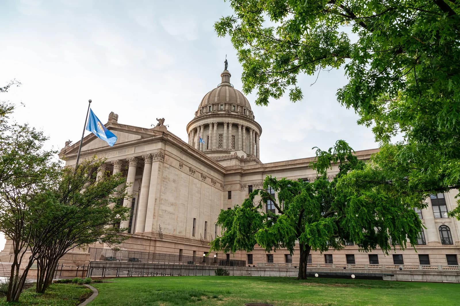 A large, light-colored stone building with a prominent dome and columns is seen from a low angle, framed by green trees and a grassy lawn. A blue flag flies from a flagpole on the left side of the building.