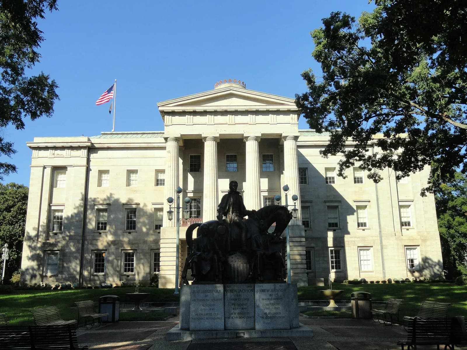 A large, light-colored stone building with a classical facade, including columns and a pediment, stands under a clear blue sky. In the foreground, a bronze equestrian statue on a stone pedestal is visible, with green lawns and trees on either side.