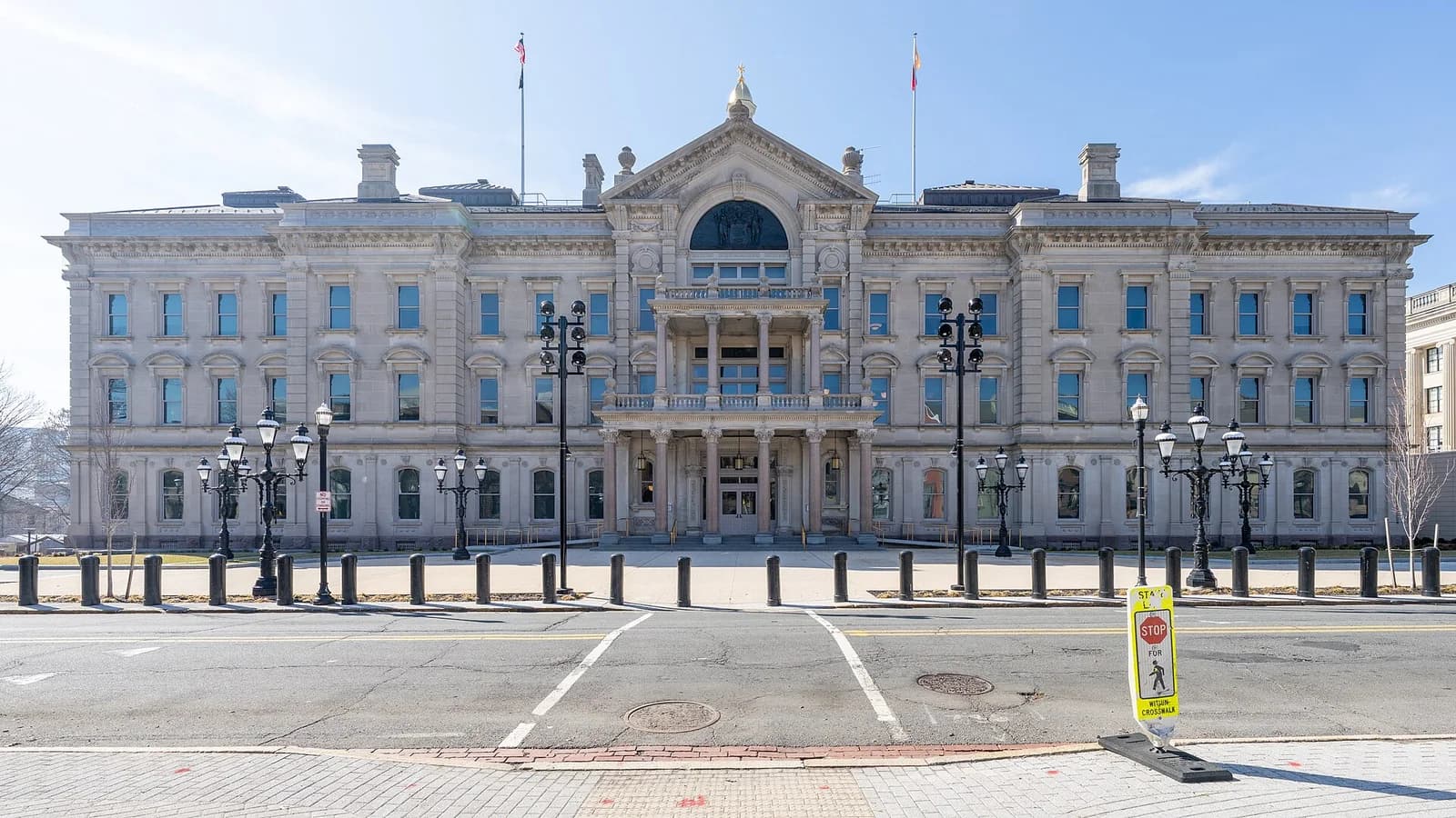 A large, ornate stone building with a central portico and columns stands behind a street with bollards and lampposts. The building has multiple windows and flags flying from its roof.