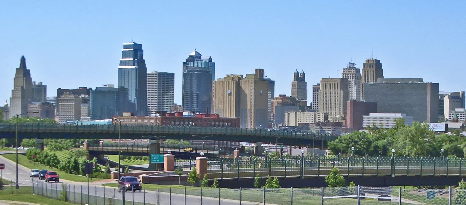 A wide shot of a city skyline with numerous tall buildings under a clear blue sky. In the foreground, a multi-level road system with cars and green landscaping is visible.