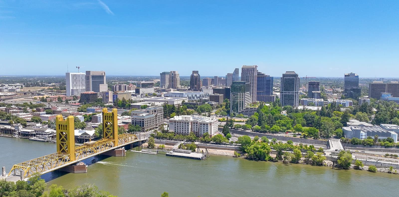 An aerial view of a city skyline features a prominent yellow bridge spanning a wide river in the foreground. Numerous high-rise buildings and green trees line the riverbanks under a clear blue sky.