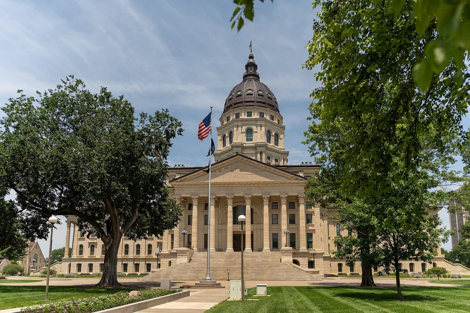 A large, light-colored stone building with a prominent dome and columns stands under a partly cloudy sky. An American flag flies on a flagpole in front of the building, which is surrounded by green lawns and trees.