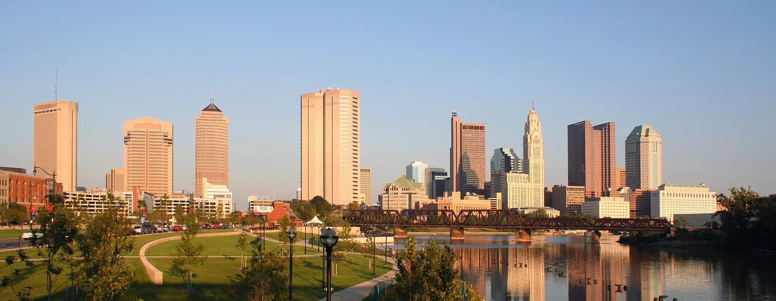 A panoramic view of a city skyline with numerous tall buildings under a clear sky. A river with reflections of the buildings flows in the foreground, with a bridge spanning across it and a green park area on the left bank.