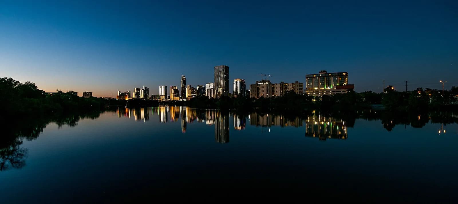 A panoramic night shot shows a city skyline with illuminated buildings reflecting on a calm body of water. The sky is a deep blue, transitioning to a lighter hue near the horizon.