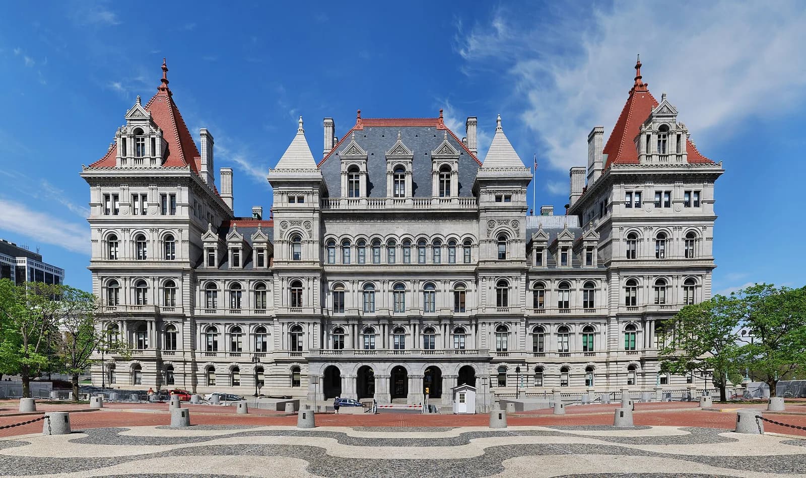A large, ornate stone building with a gray and red roof stands under a clear blue sky. The building features multiple windows, turrets, and intricate architectural details, with a patterned plaza in the foreground.