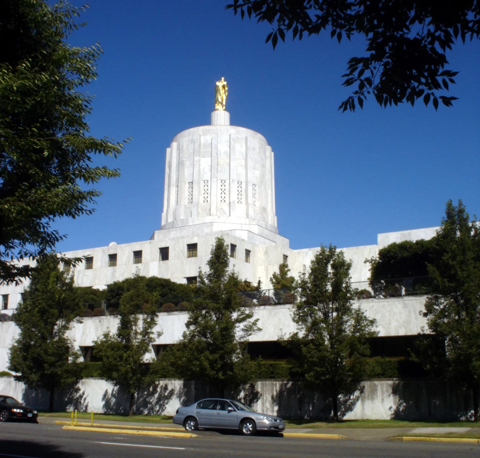 A white marble building with a large dome topped by a golden statue stands against a clear blue sky. Trees frame the building, and a street with parked cars is visible in the foreground.
