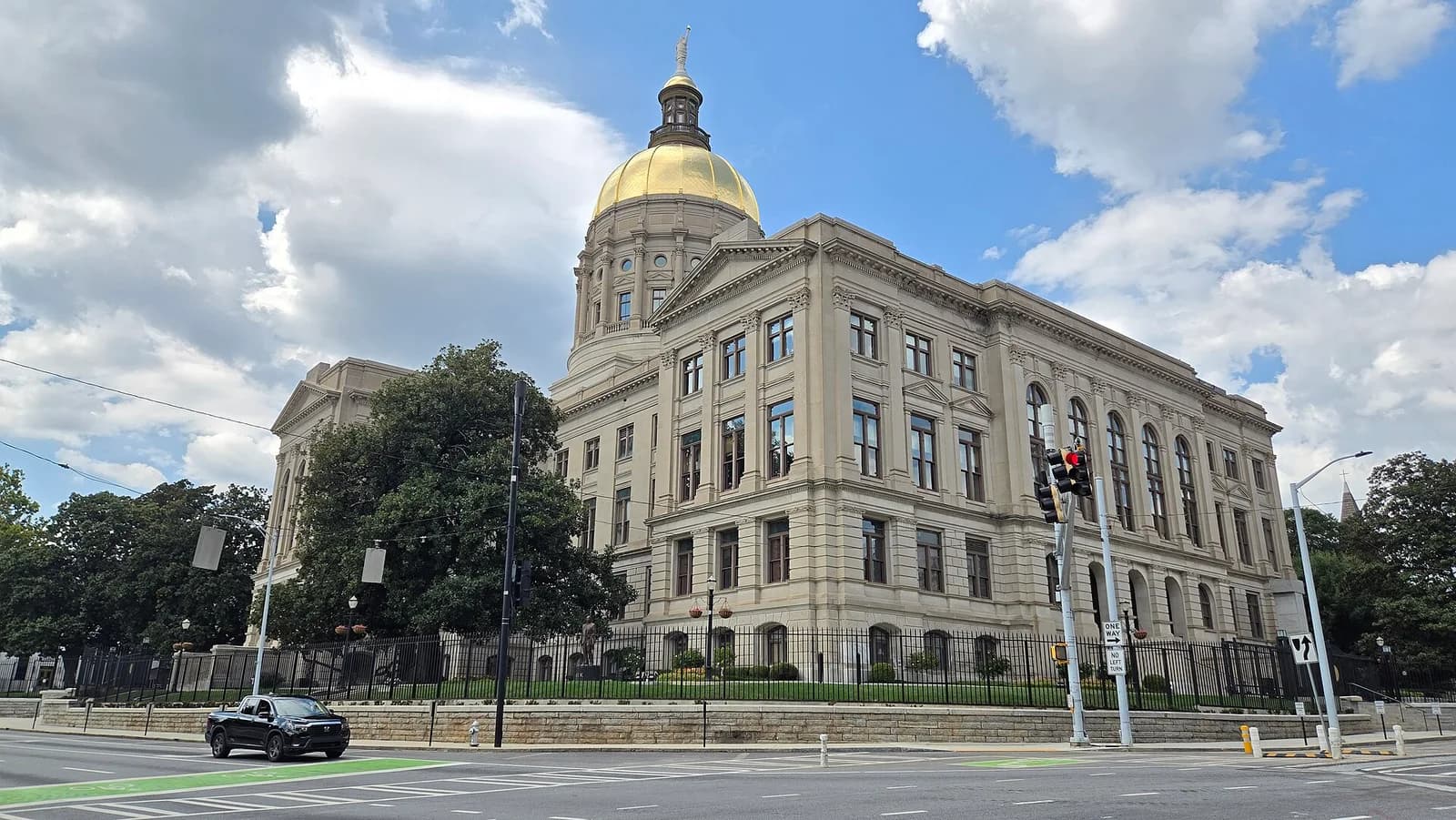 A large, ornate building with a prominent golden dome stands under a partly cloudy sky. A black car drives on a street in the foreground, with traffic lights and street signs visible.