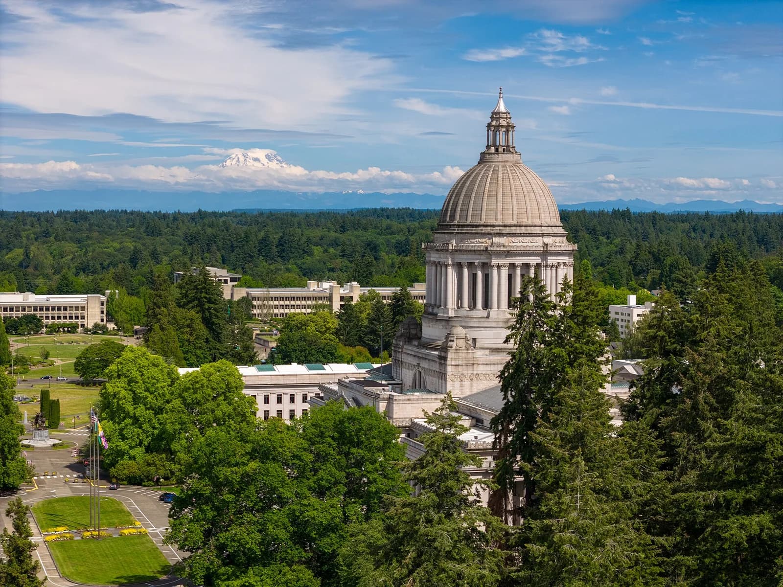 An aerial view shows a large domed building with columns, surrounded by green trees and other institutional buildings. In the far distance, a snow-capped mountain rises above a forested landscape under a partly cloudy sky.