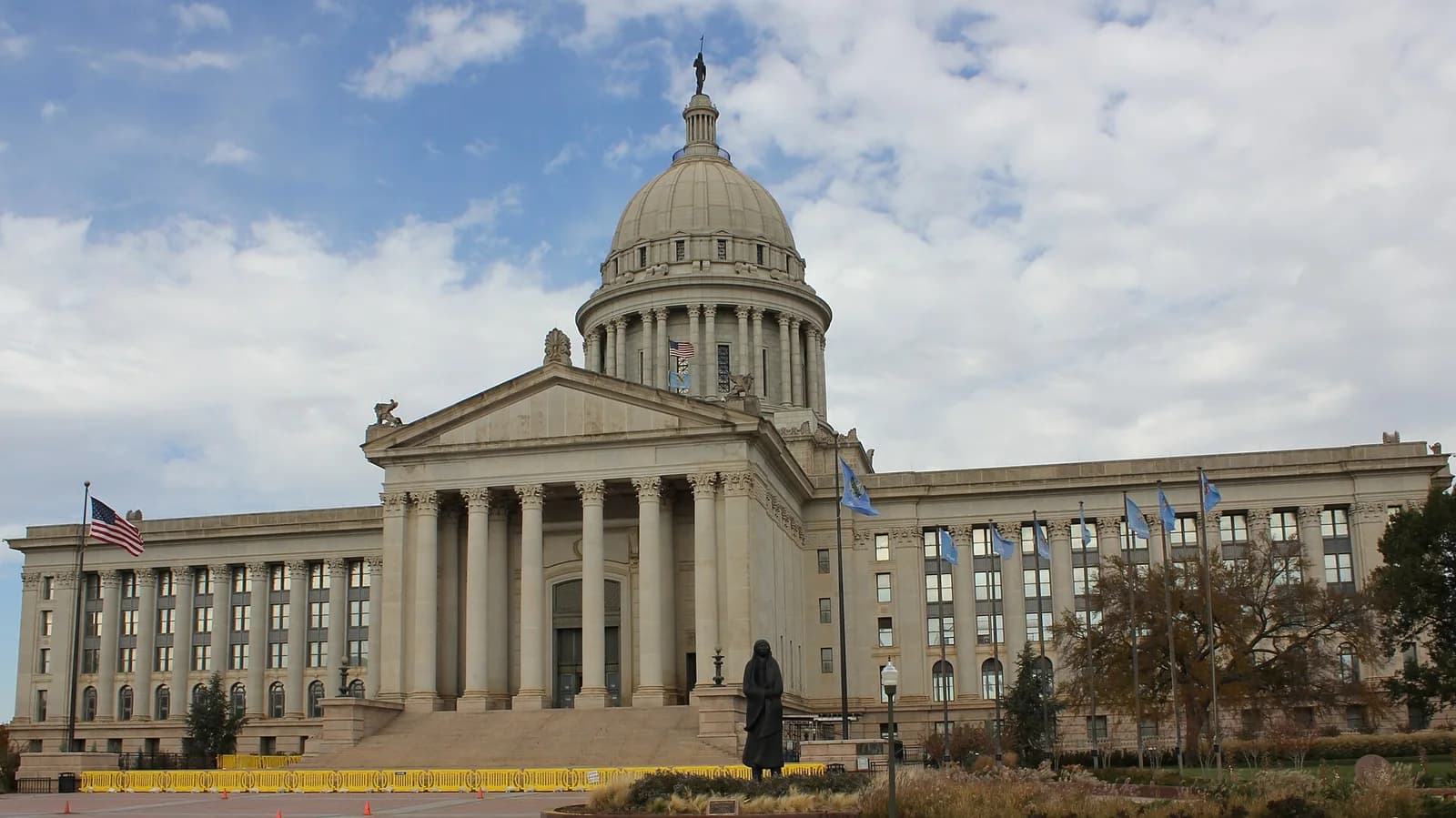 A large, classical-style building with a central dome and a grand portico with columns is shown under a partly cloudy sky. An American flag flies on the left, and several light blue flags are visible on the right, with a statue in the foreground.