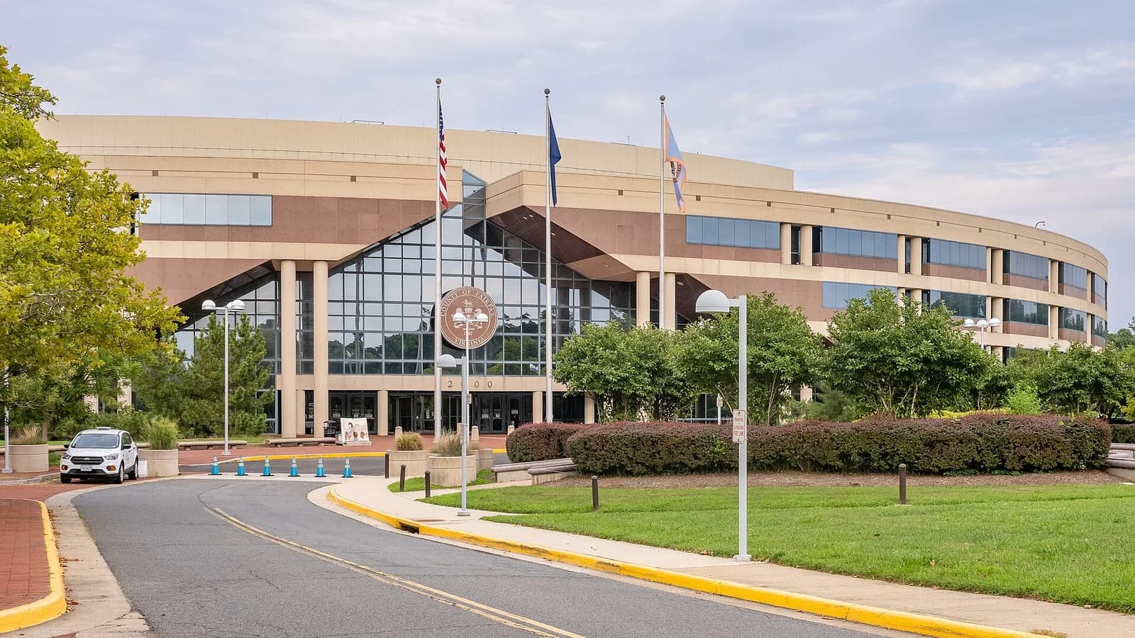A modern, multi-story building with a curved facade and a large glass entrance is visible from a paved driveway. Three flagpoles with flags stand in front of the entrance, and landscaping with trees and shrubs surrounds the building.