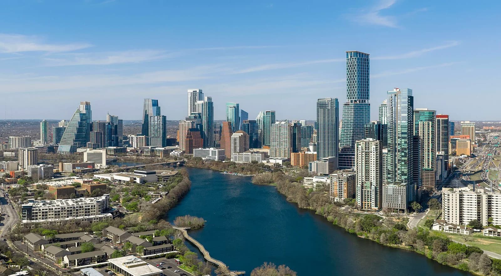 An aerial view of a city skyline with numerous tall buildings, some with distinctive architectural designs, lining the banks of a wide river. The river winds through the urban landscape, with green trees and some smaller buildings visible along its edges.
