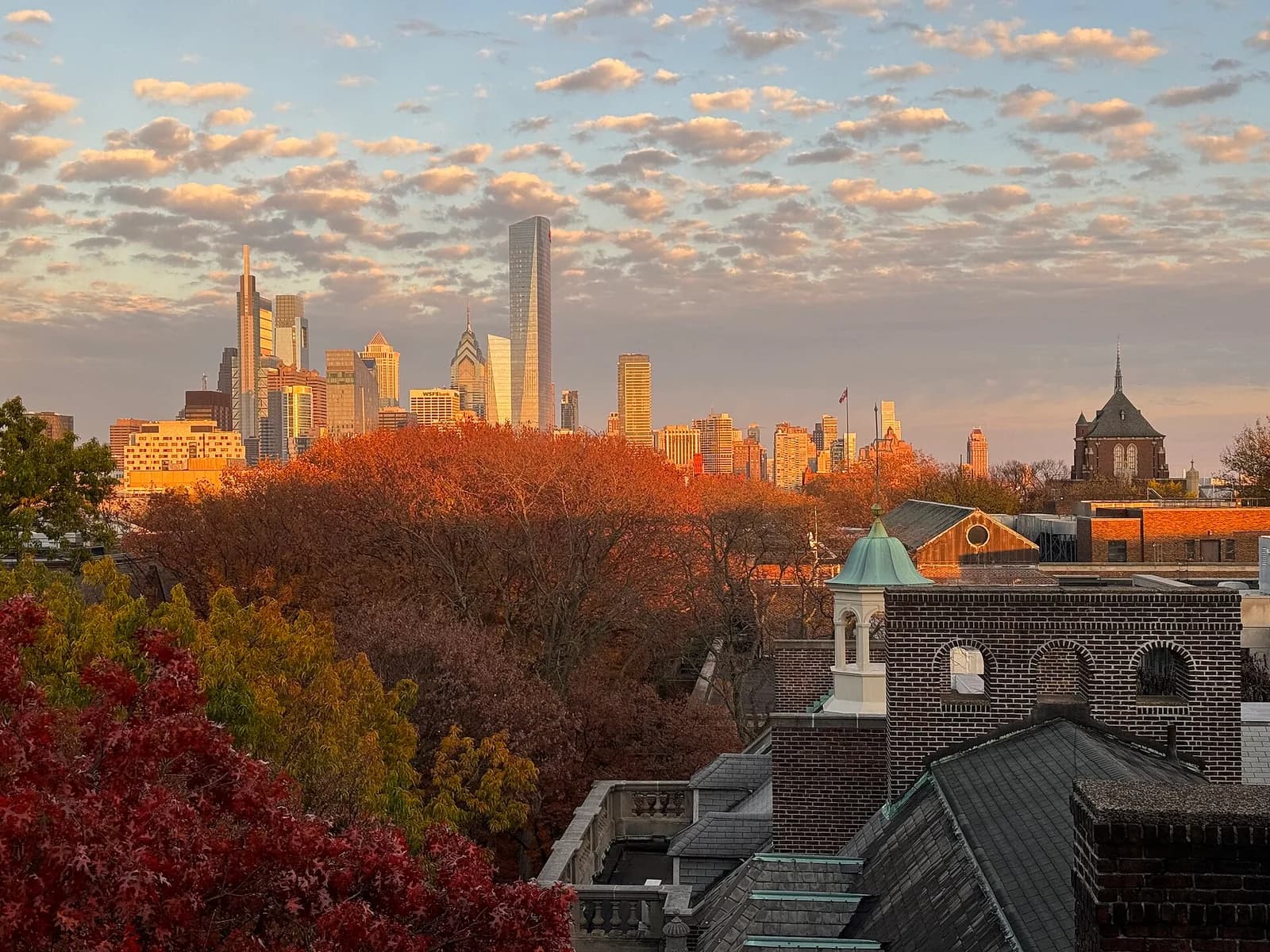 A cityscape at sunset with a skyline of tall buildings in the background, framed by autumn trees and the rooftops of foreground buildings. The sky is filled with scattered clouds, illuminated by the warm light of the setting sun.