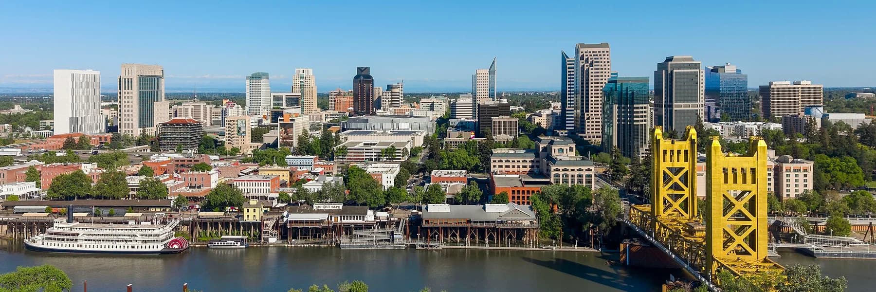 An aerial panoramic view shows a city skyline with numerous buildings of varying heights, a river in the foreground with a paddlewheel boat docked, and a prominent yellow bridge spanning the river on the right.