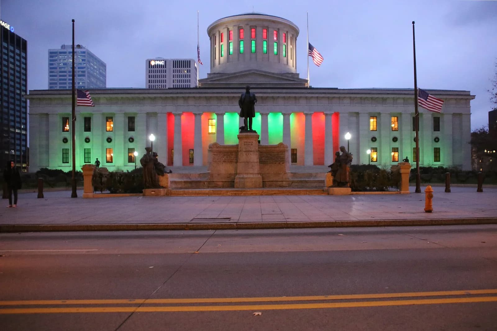 A large, classical-style building with a central dome is illuminated with red and green lights. In front of the building is a statue on a pedestal, flanked by two smaller statues, with a street in the foreground.