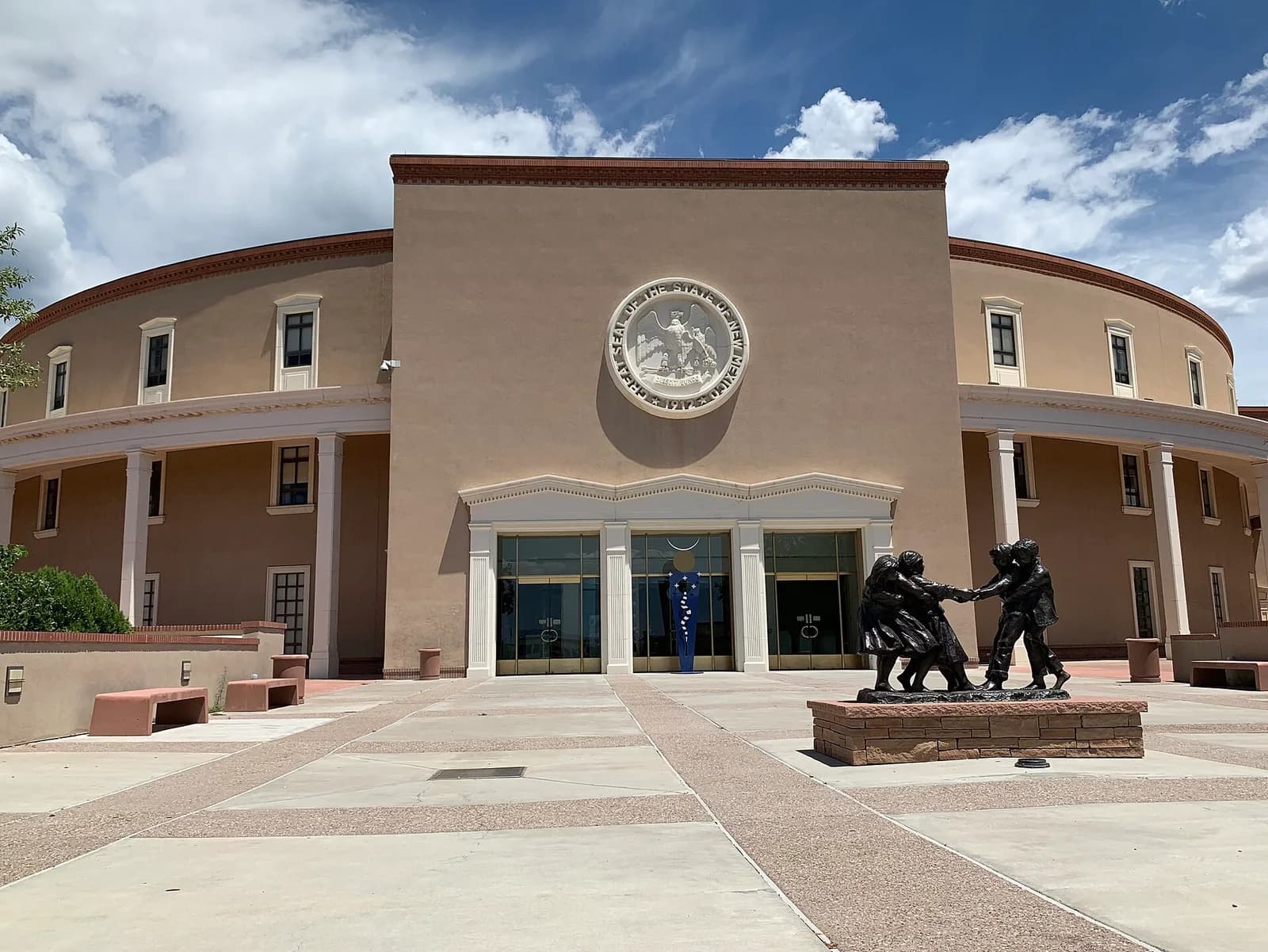 A tan-colored institutional building with a circular facade and a large seal above the main entrance is shown under a partly cloudy sky. In front of the building, a bronze sculpture of three figures stands on a brick base.