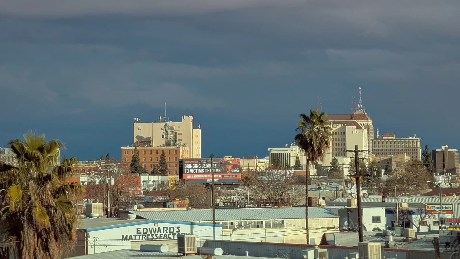 A cityscape under a dark, cloudy sky shows various buildings, including a prominent billboard with text and several palm trees in the foreground. The buildings range from multi-story structures to smaller commercial buildings with flat roofs.