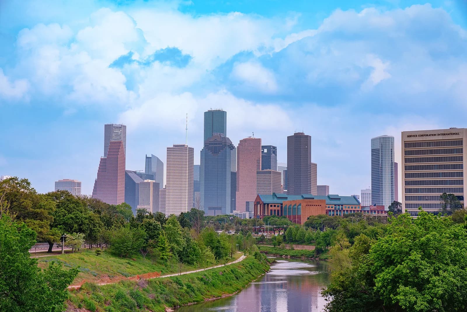 A cityscape features numerous tall buildings under a blue sky with white clouds. In the foreground, a river reflects the sky, bordered by green trees and a grassy bank with a walking path.