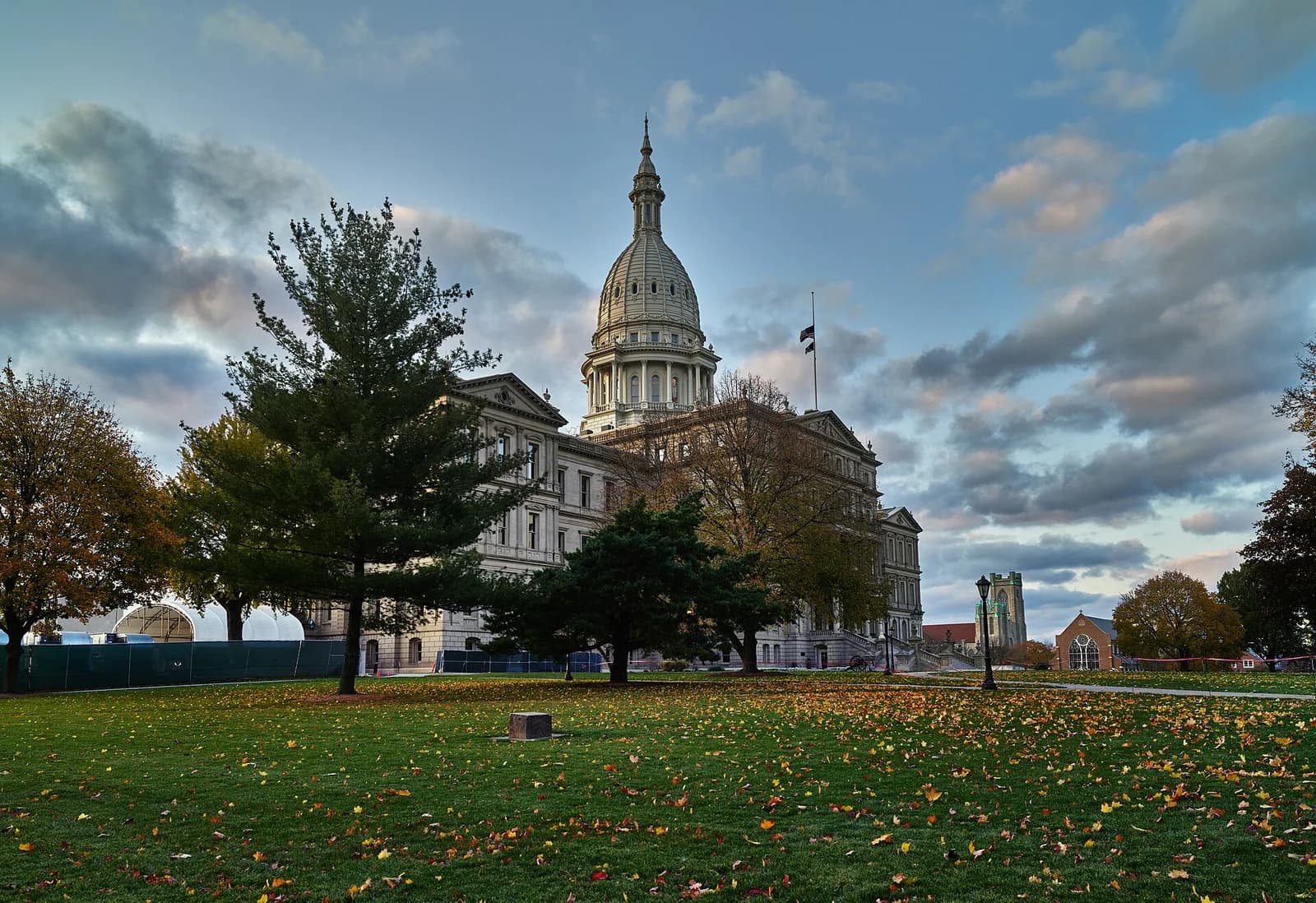 A large, light-colored institutional building with a prominent dome and spire stands under a cloudy sky. In the foreground, a green lawn is scattered with autumn leaves, and several trees frame the building.