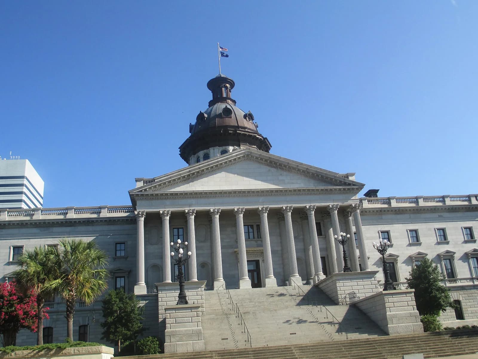 A large, light-colored stone building with a prominent dome and columns is shown under a clear blue sky. A flag flies from the top of the dome, and a modern building is visible in the background to the left.