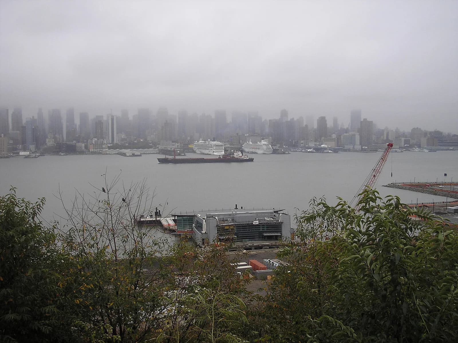 A cityscape view on a foggy day shows a body of water with several ships and a distant skyline of tall buildings partially obscured by mist. In the foreground, green trees and a modern building with a dock are visible, along with a red construction crane.