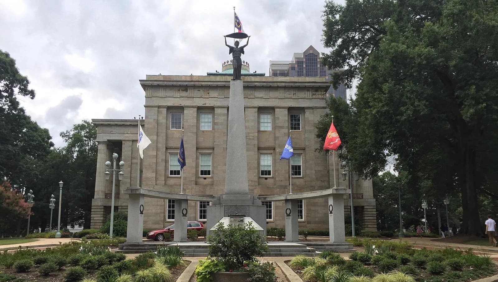 A large, light-colored stone building with columns and multiple windows stands behind a tall monument topped with a statue holding a flag. Several other flags fly on poles around the monument, and green trees and landscaping surround the area under a cloudy sky.