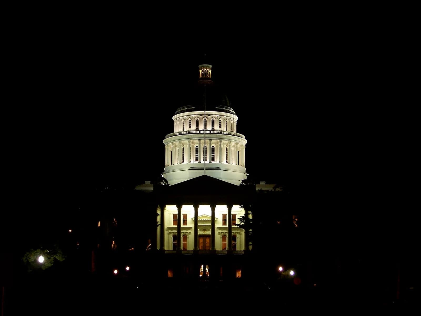 A grand, white domed building is brightly illuminated at night against a dark sky. The building features a large portico with columns and a multi-tiered dome topped with a cupola.