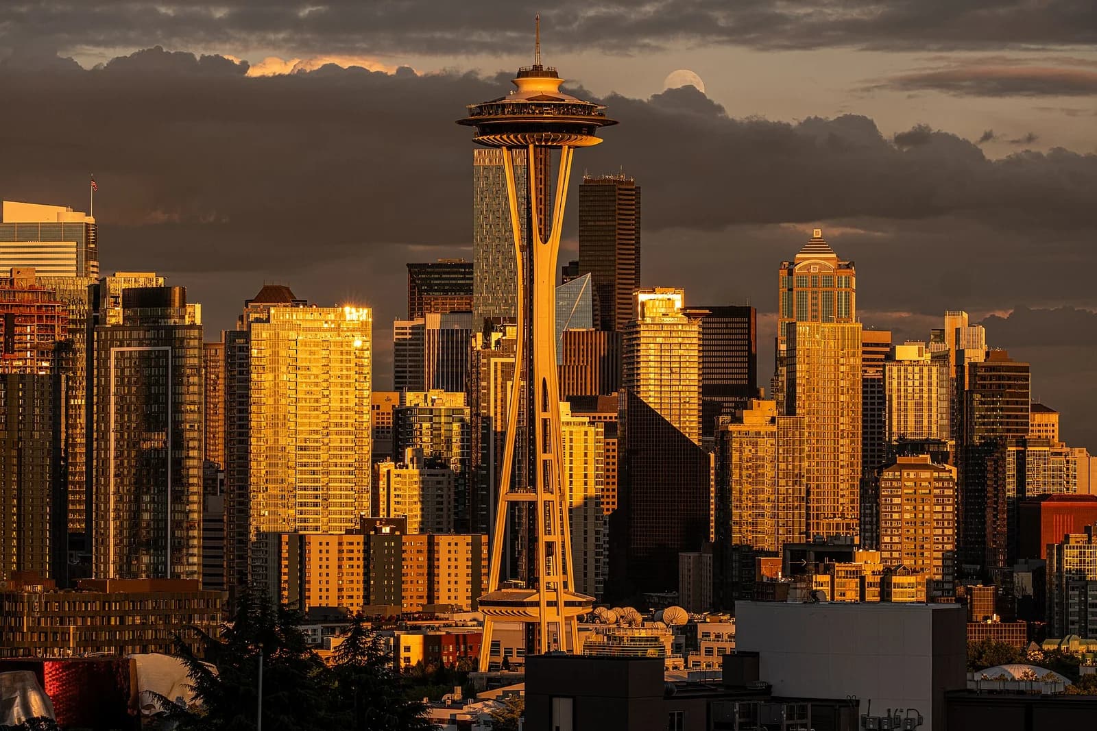 A cityscape at sunset shows a tall, slender tower with a circular top, surrounded by numerous high-rise buildings. The setting sun casts a golden glow on the buildings, and a full moon is visible in the cloudy sky above.